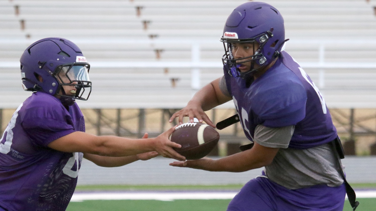 Two football players in purple uniforms perform a handoff during practice on a field with bleachers in the background.