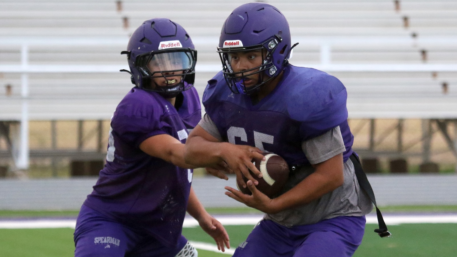 Two purple-uniformed football players in helmets contest for the ball during practice on a field, one holding the ball while the other reaches in.