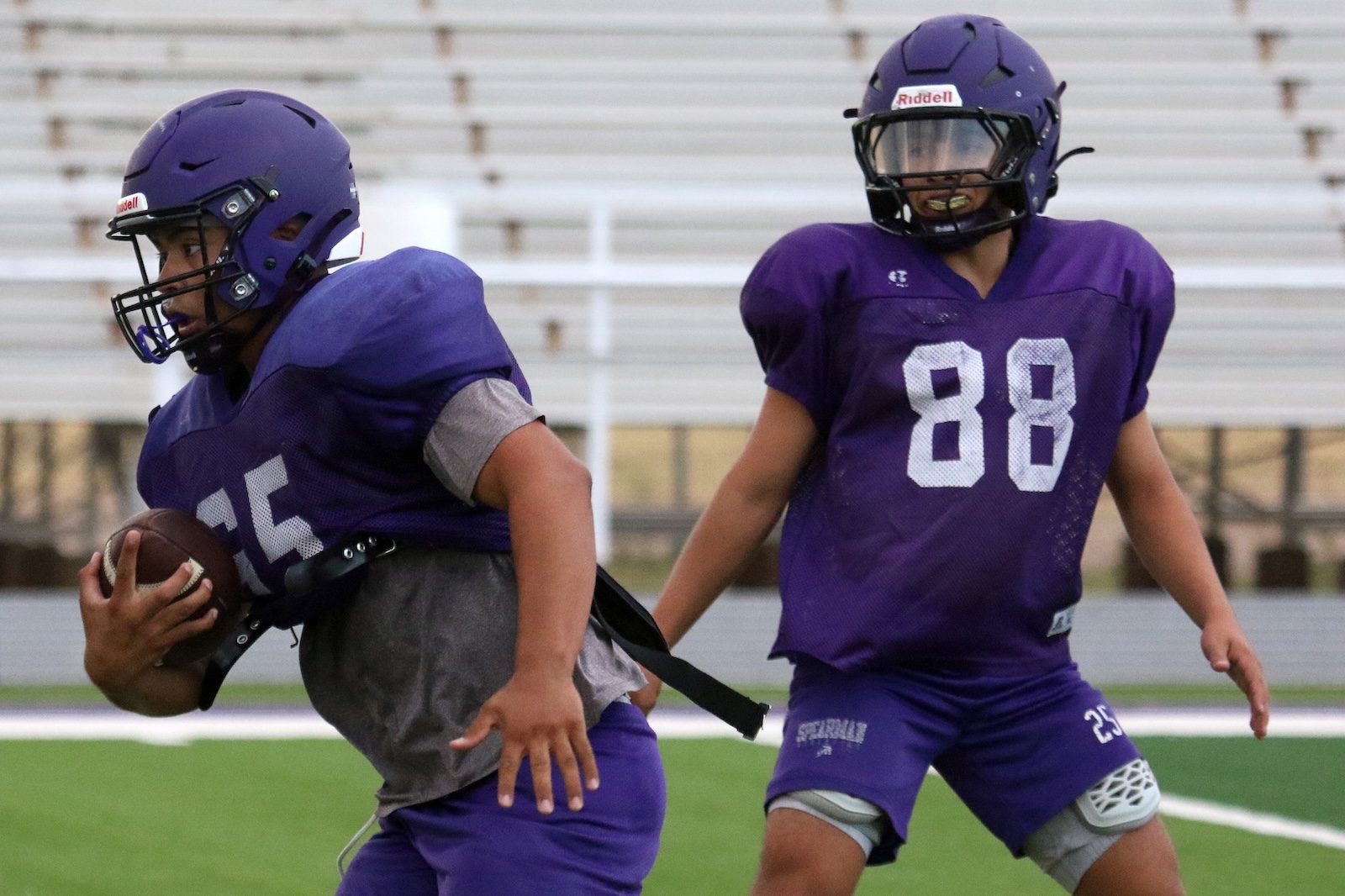 Two football players in purple uniforms on a field; one (#15) runs with the ball, the other (#88) moves in to support/defend.