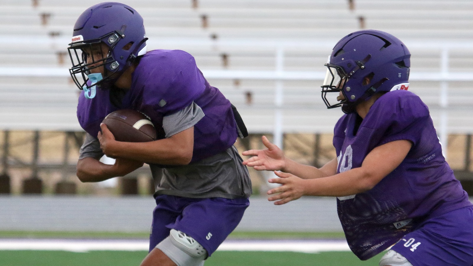 Two football players in purple uniforms and helmets; one carries the ball as a teammate closes in.
