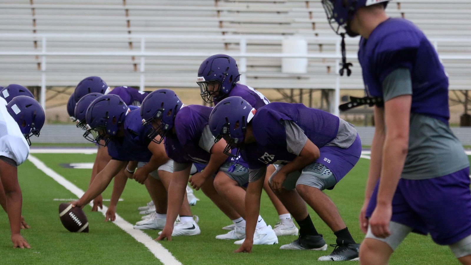 Football team lines up at the line of scrimmage in purple jerseys, preparing for snap on a green field.