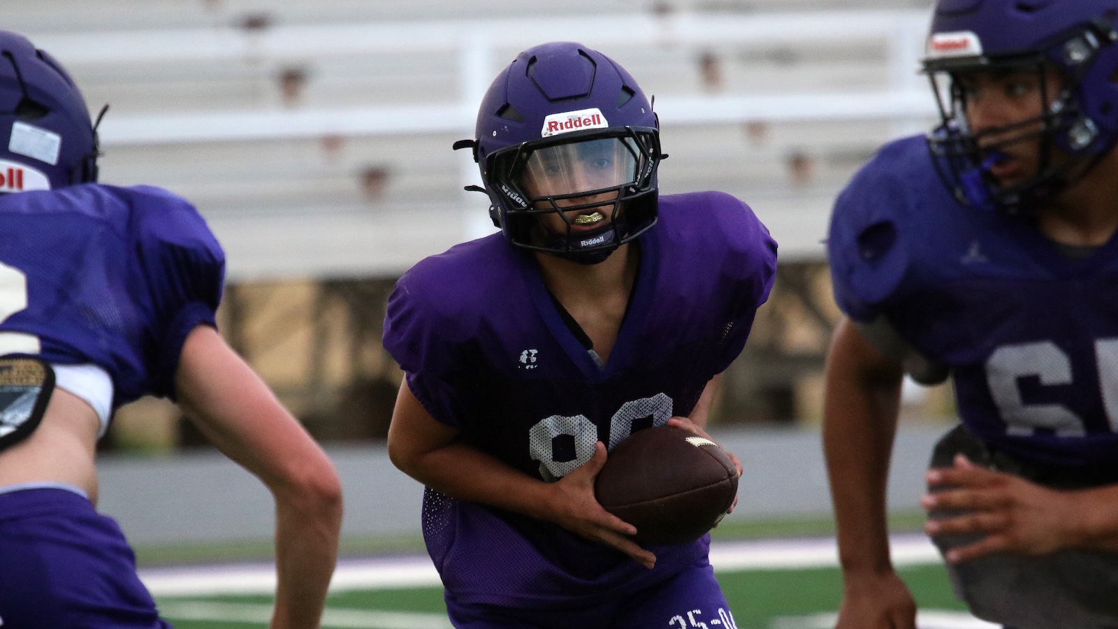 Young football player in a purple uniform holding a football, wearing a helmet, ready to run on the field.