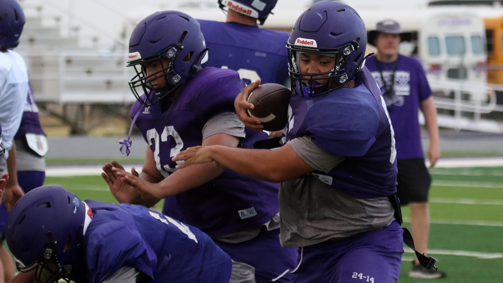 Young football players in purple uniforms tackle on a field; one holds a ball while others push forward.