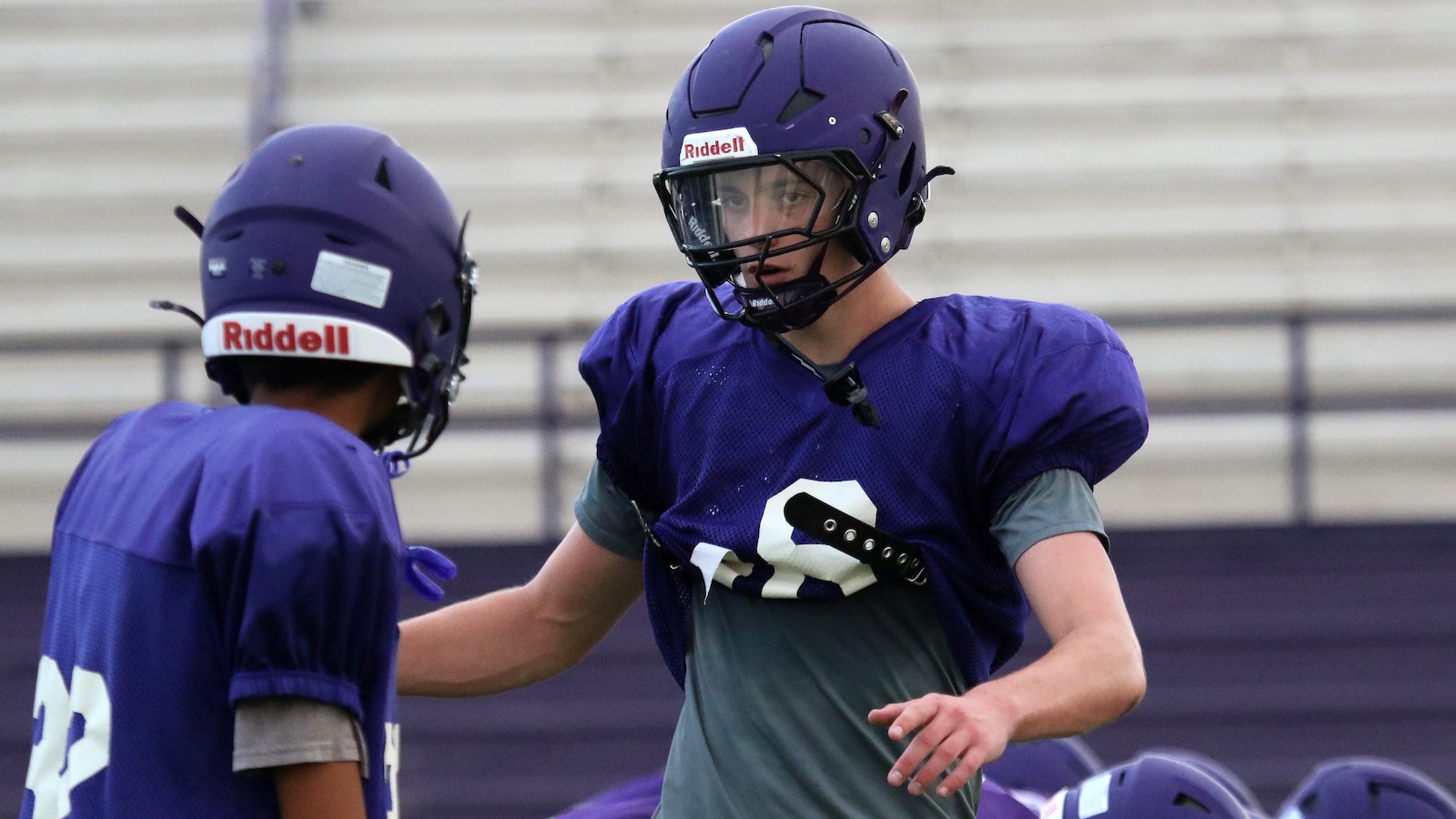 Two football players in purple helmets and jerseys practice on a field, one facing the camera with a protective visor while the other talks nearby.