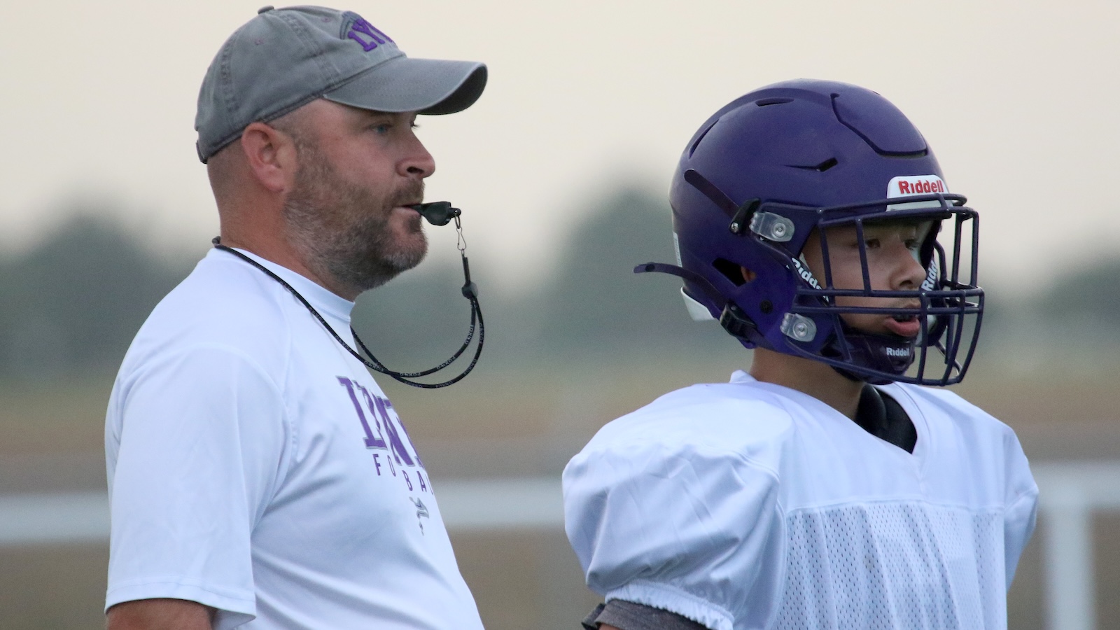 Coach with whistle watching a young football player in a purple helmet during practice.