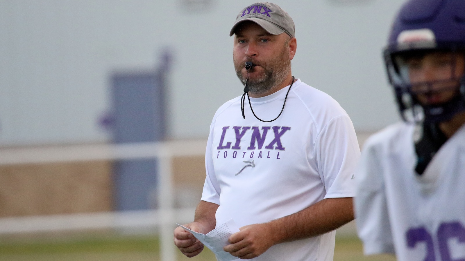 Football coach in a white Lynx Football shirt and gray cap, whistle in mouth, holding a play sheet on a field.