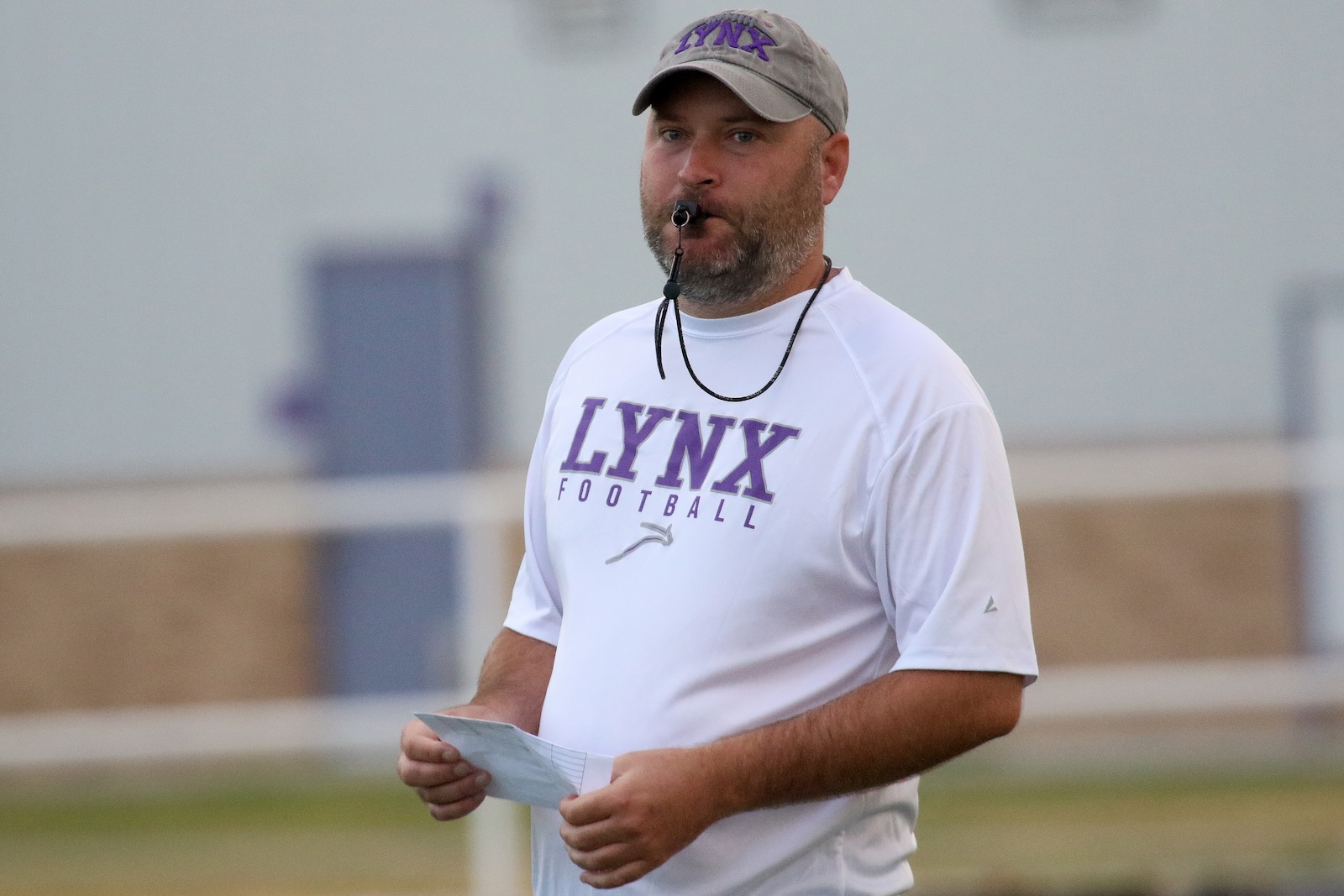 Male football coach wearing a white LYNX Football shirt, with a whistle in his mouth, holding a note sheet on a field.