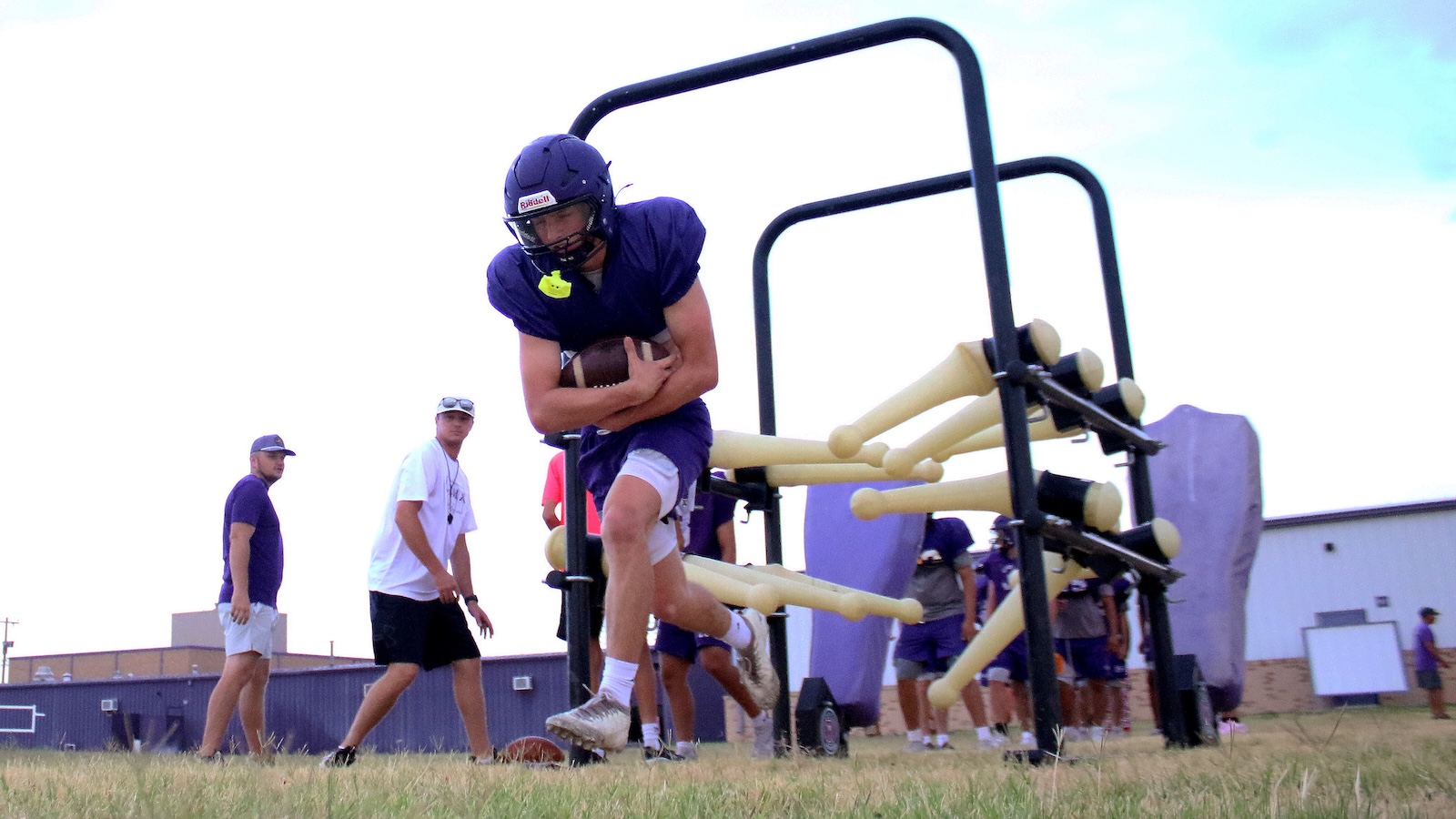 Football player in purple uniform sprints while cradling a ball through a hurdle-style sled during practice with teammates watching in the background.
