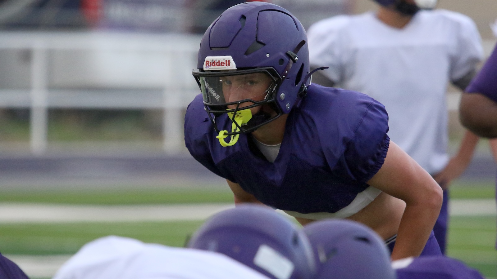 Football player in purple helmet and jersey crouches in a ready stance during practice; neon yellow mouthguard visible.