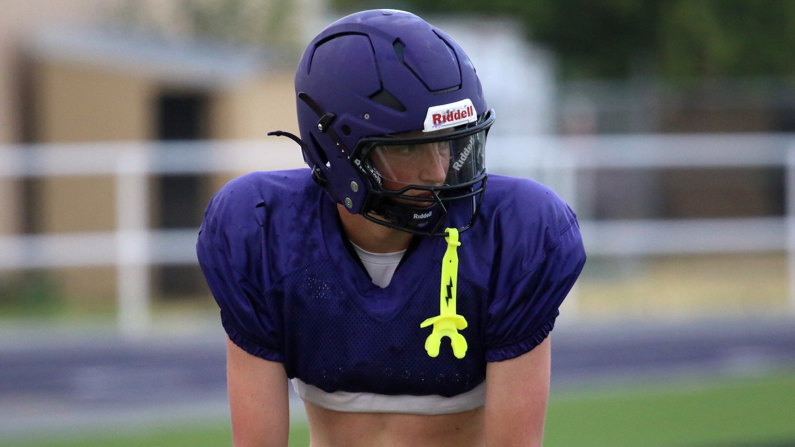Football player in a purple helmet and jersey on a practice field, with a neon yellow flag belt hanging from the chest helmet area.