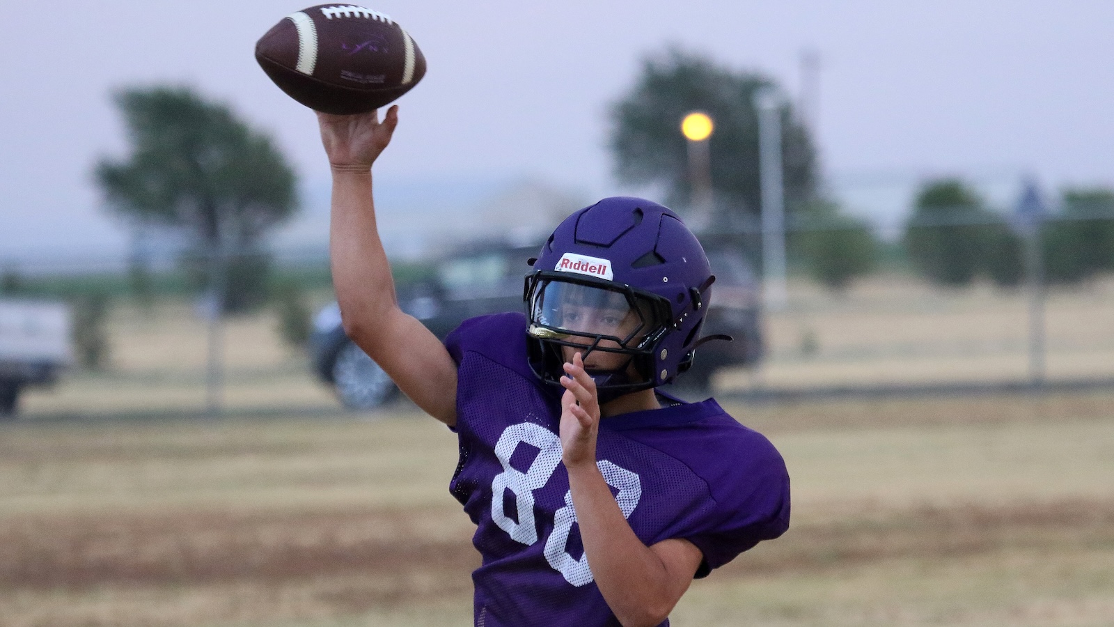 Youth football player in a purple jersey and helmet throws a football on a grassy field during practice