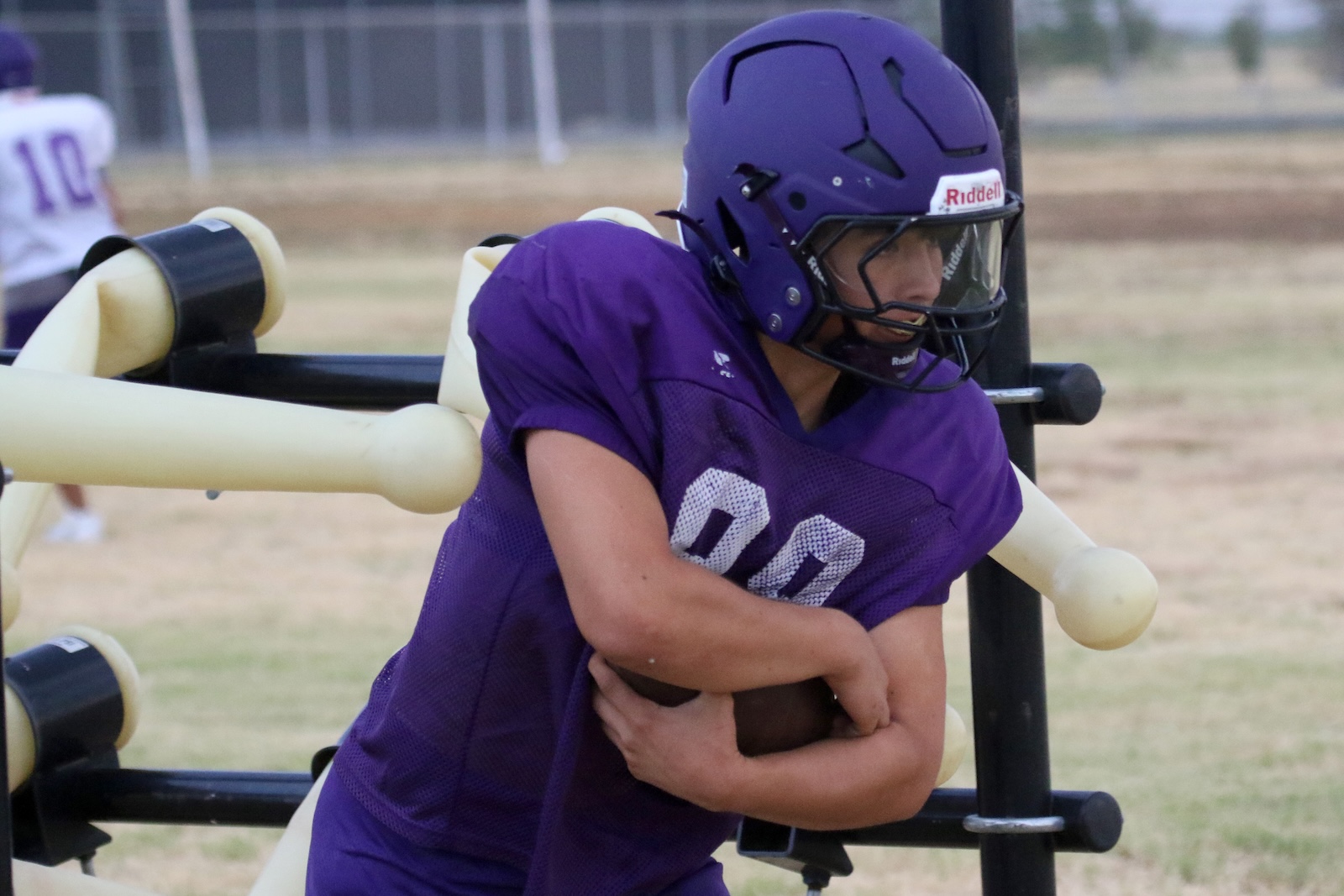 Football player in a purple uniform and helmet cradling a ball during practice, leaning into a fake blocker structure on the field.
