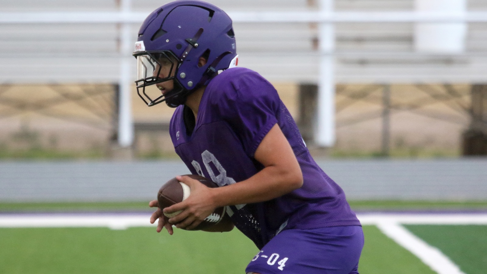 Football player in a purple jersey and helmet, holding a football and sprinting on a sideline field to make a play.