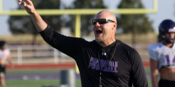 Coach on a football field raising his arm and pointing, wearing a black long-sleeve shirt and sunglasses.