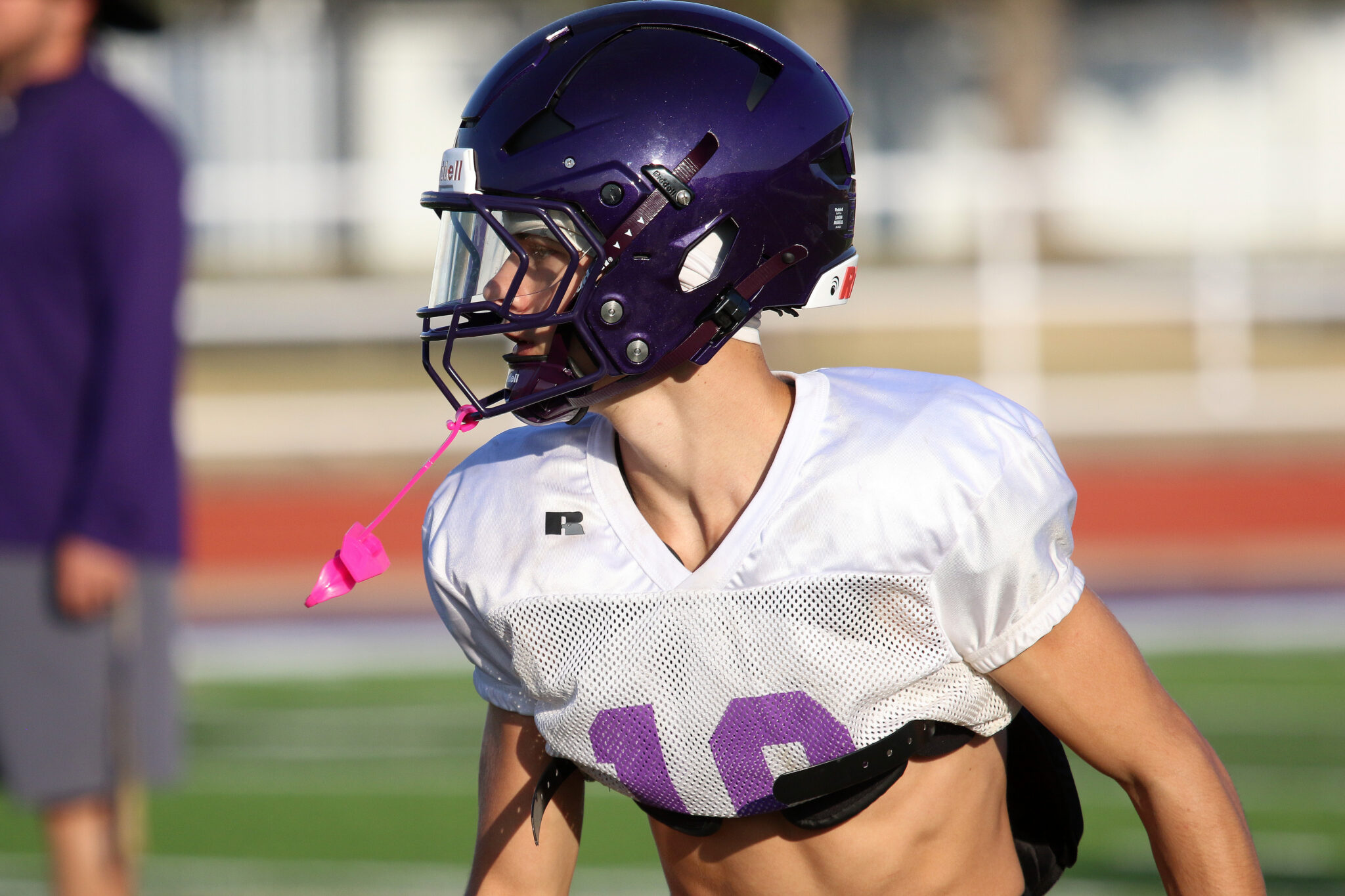 1K6A1327 | Press Pass Sports Football player in a purple helmet and white jersey with a purple number, on a football field with a pink mouthguard hanging from the gear.