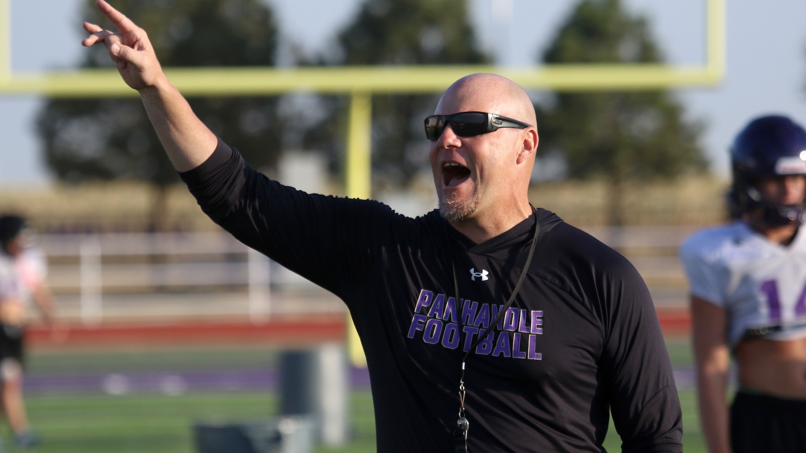 Bald man wearing sunglasses on a football field, shouting and pointing with his arm raised during practice.