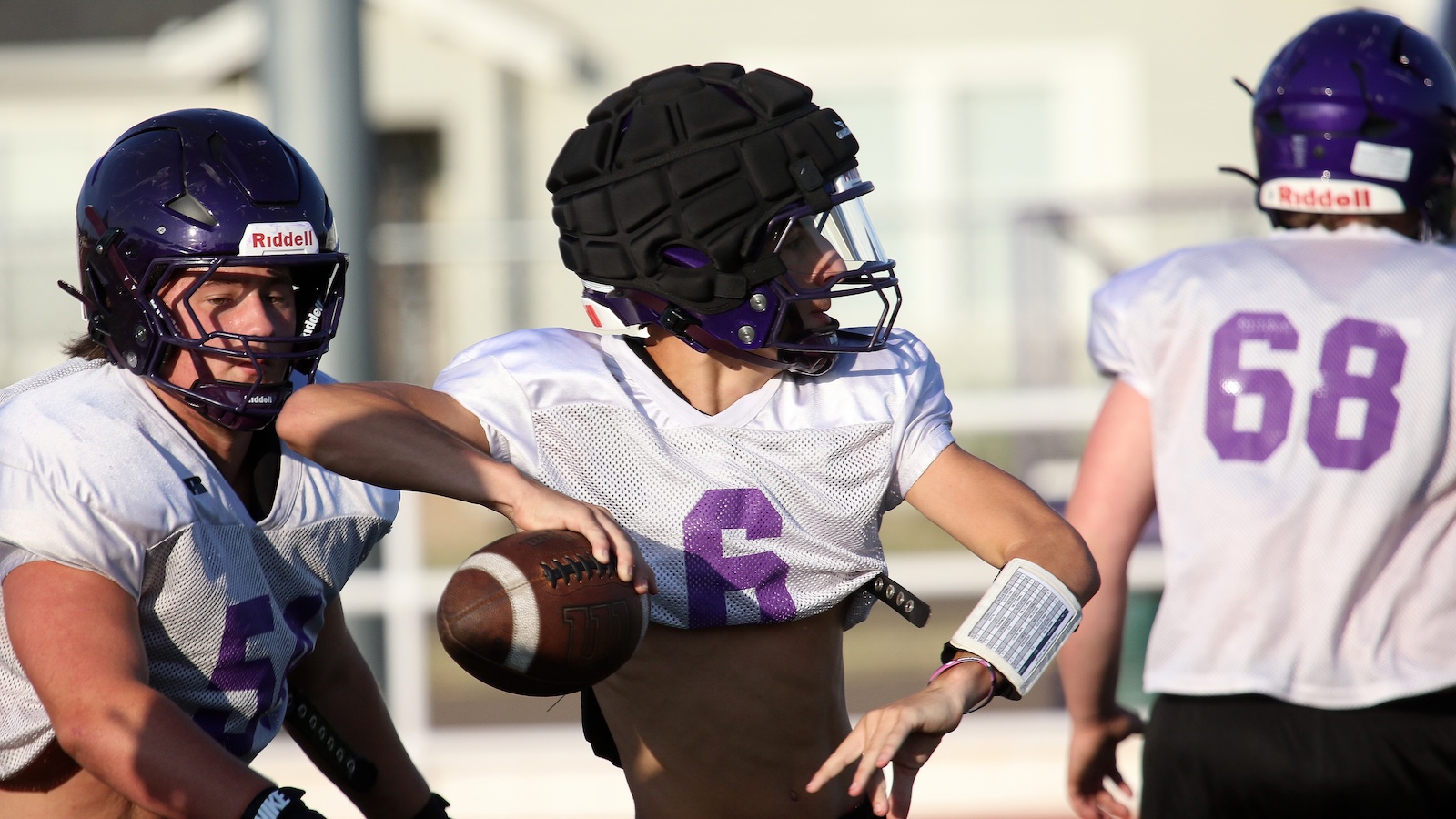 Three football players in purple and white practice jerseys during drills; central player grips a football and wears a padded helmet with face guard.
