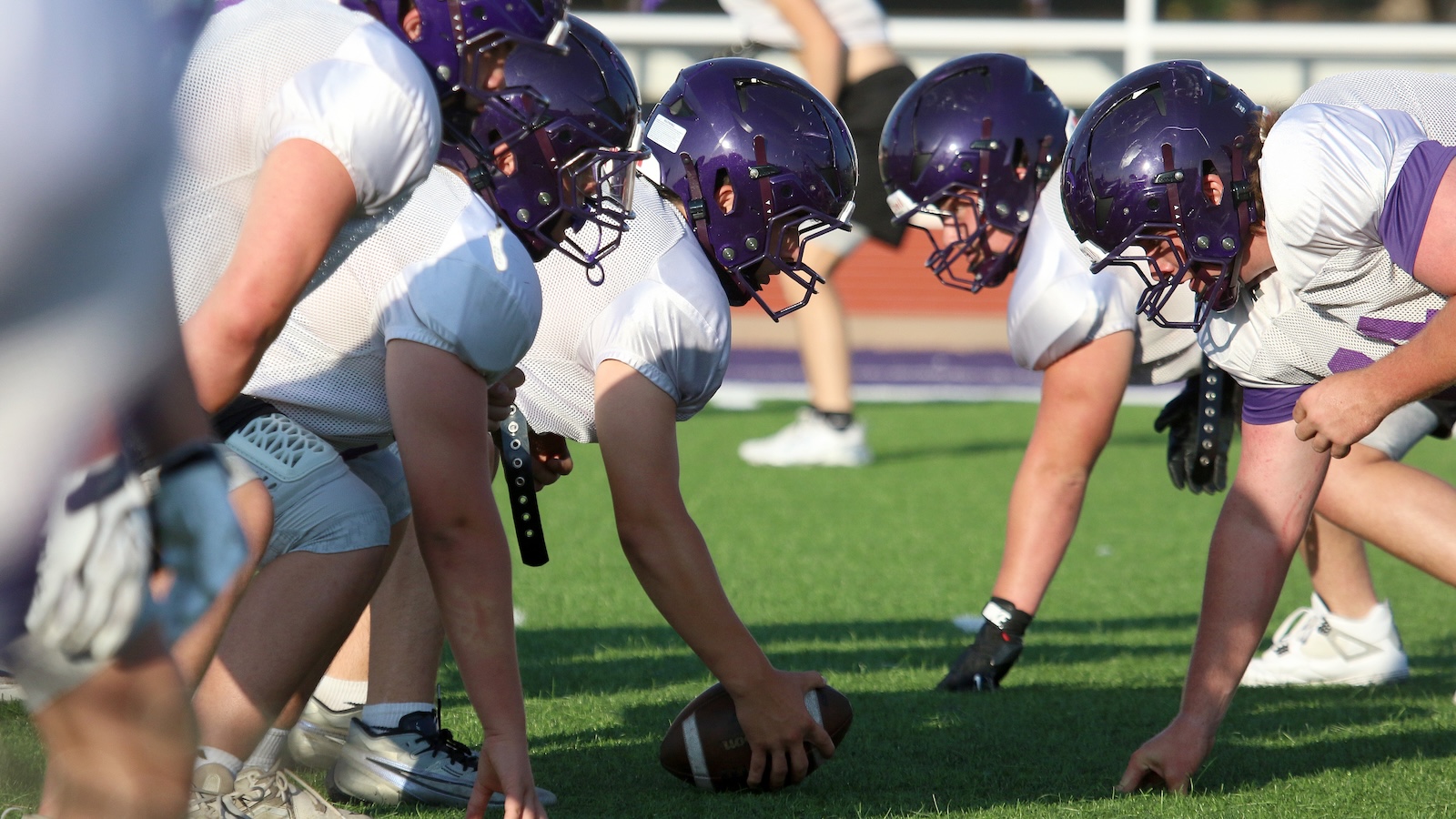 Football players in formation at the line of scrimmage, center snapping the ball to the quarterback on a green field, all wearing purple helmets and white uniforms.