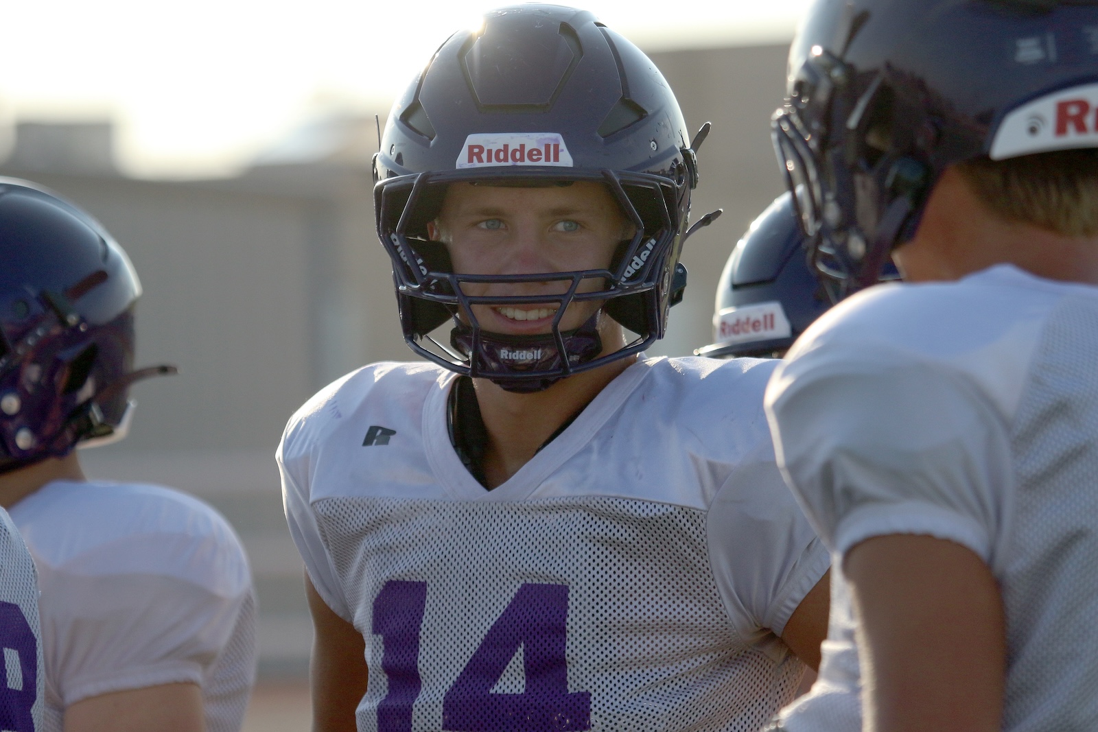 Young football player wearing a black Riddell helmet and white jersey with purple number 14, smiling at teammates during practice.