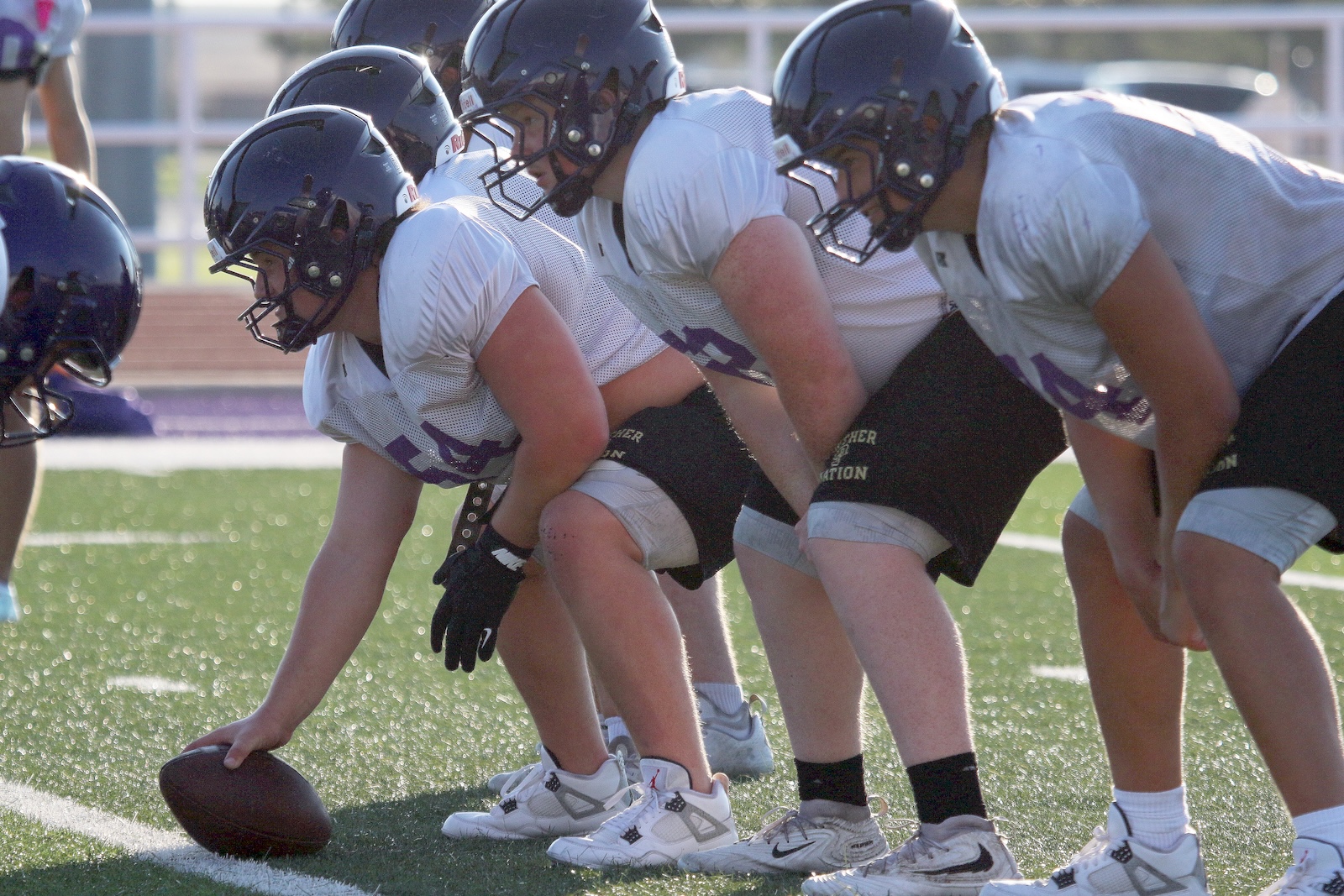 American football linemen in white jerseys crouch at the line of scrimmage, preparing to snap the ball on a sunny field.
