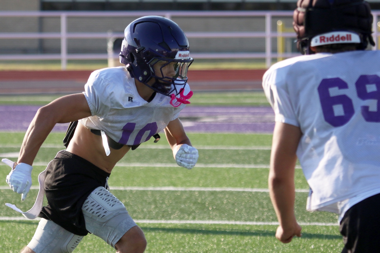 Two football players in white jerseys sprint on a green turf field during a drill, one wearing a navy helmet and pink mouthguard (#10).