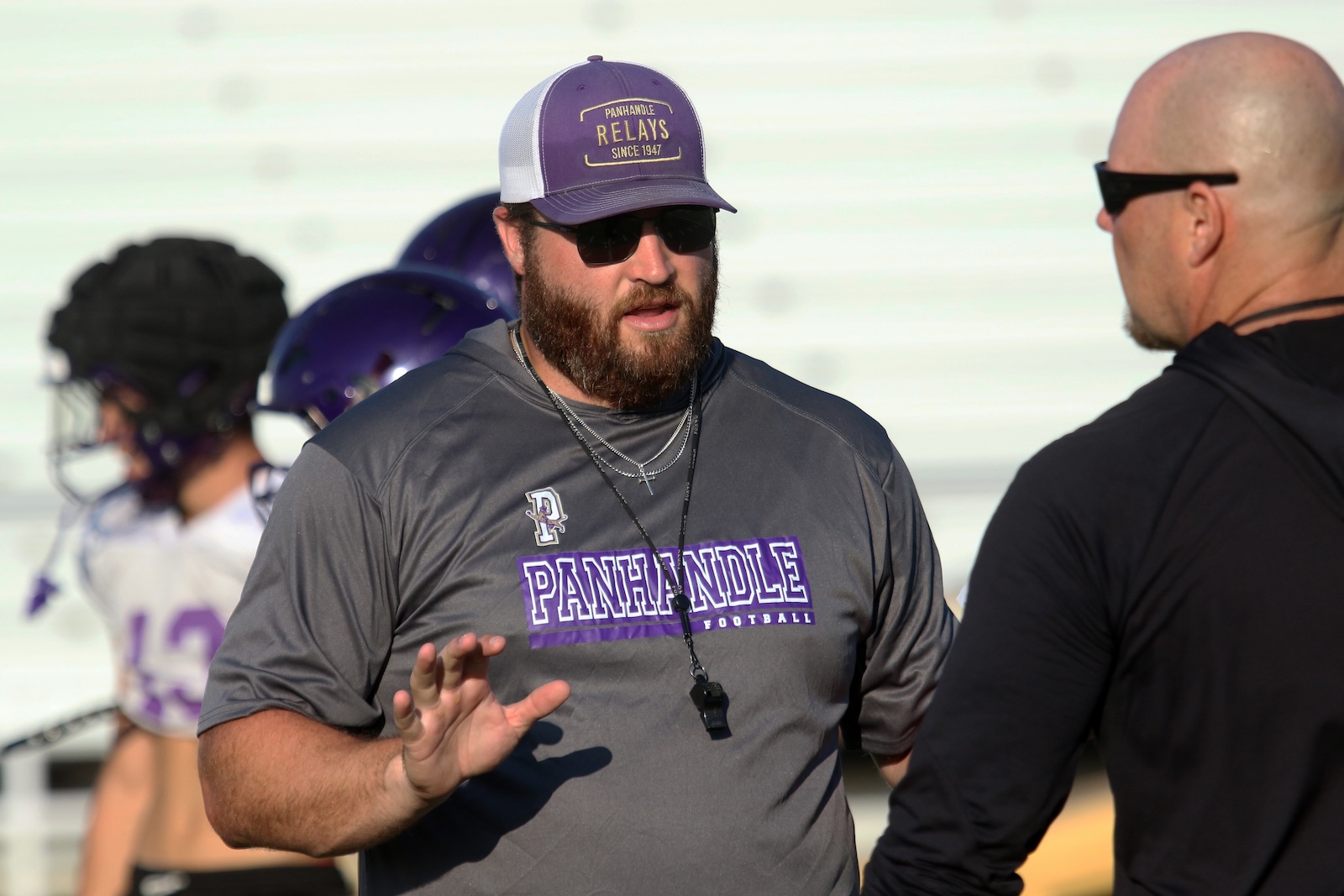 Bearded coach in a gray Panhandle Football shirt talks to a teammate on a sunny football field.