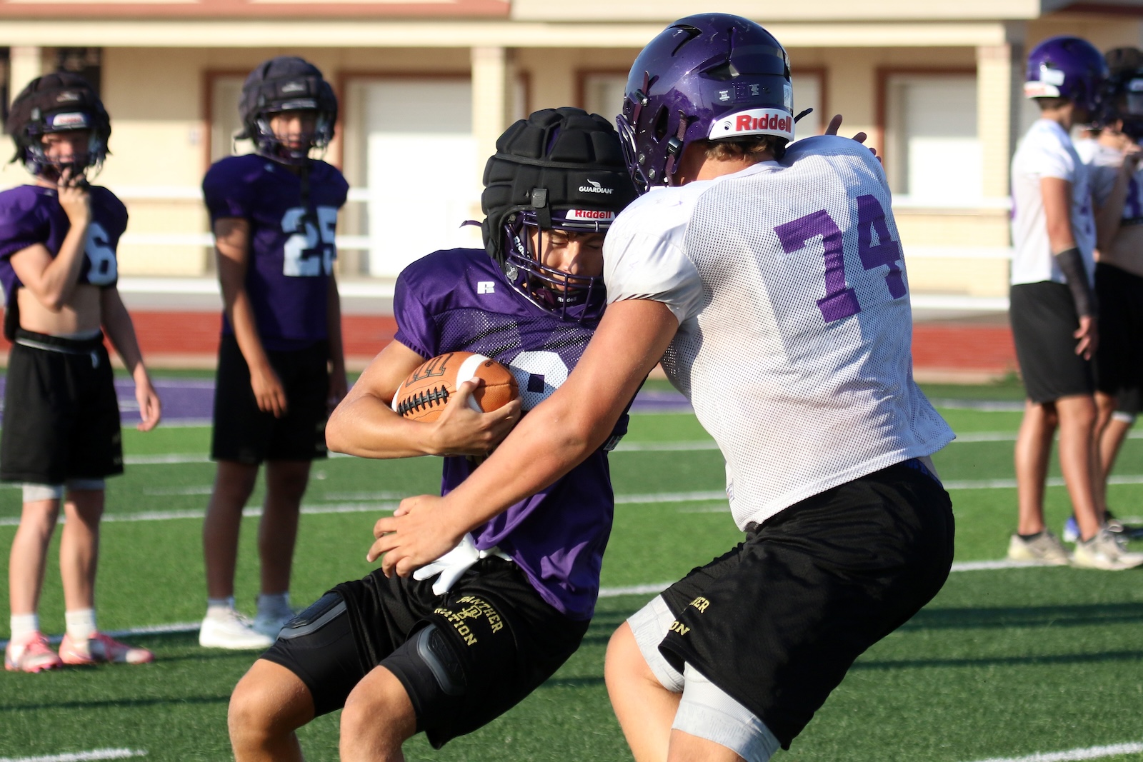 Youth football action: a purple-jersey runner protects the ball as a white-jersey defender (#74) tackles him on a sunny field.