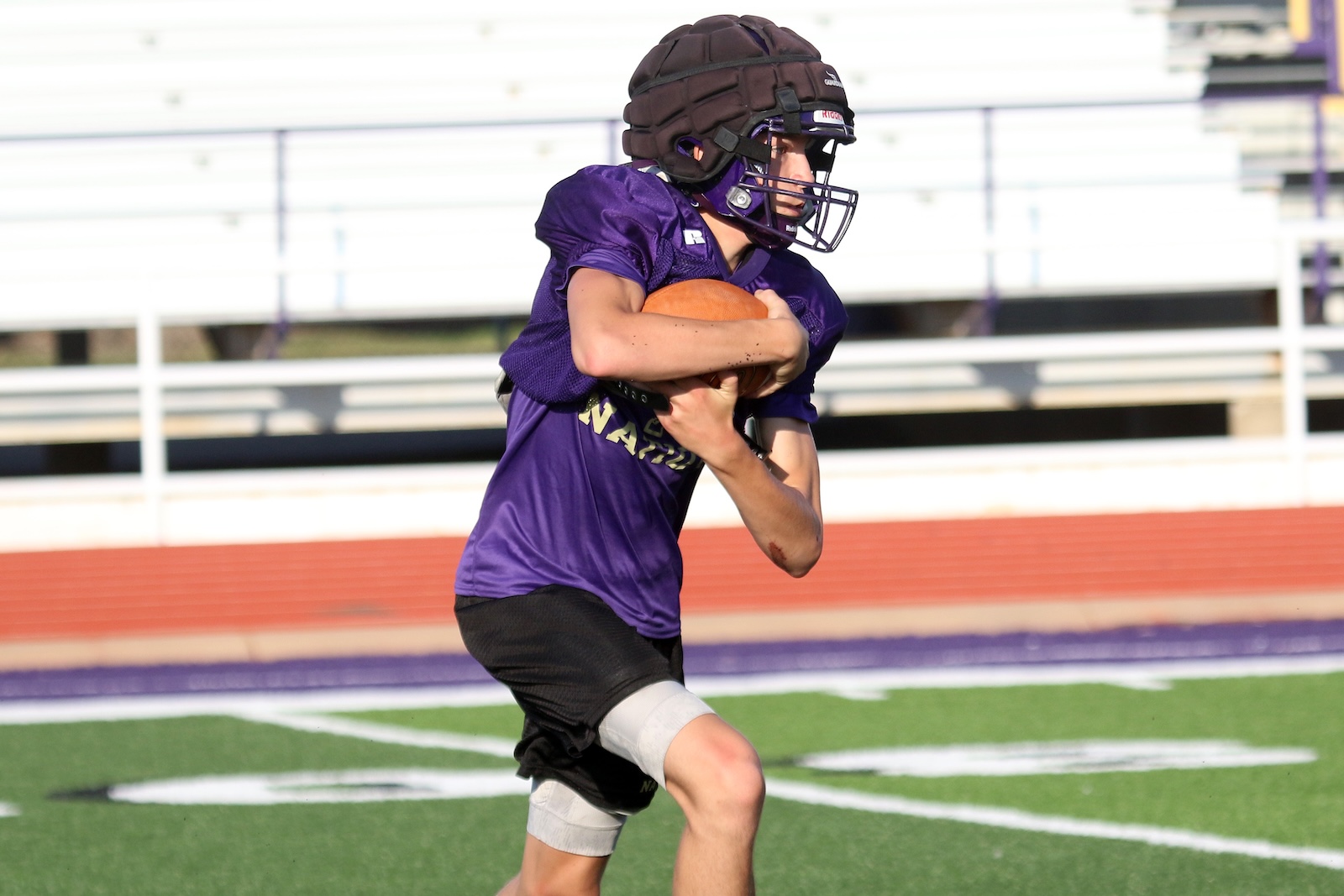Football player in a purple jersey and helmet running with a football tucked under his arm on a green turf field, stadium in background.