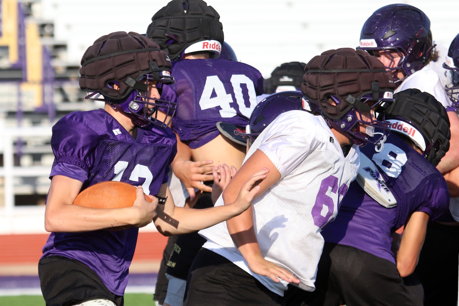 Football players in purple uniforms practice on a sunny field, with a quarterback gripping a ball as teammates block and push in a drill.