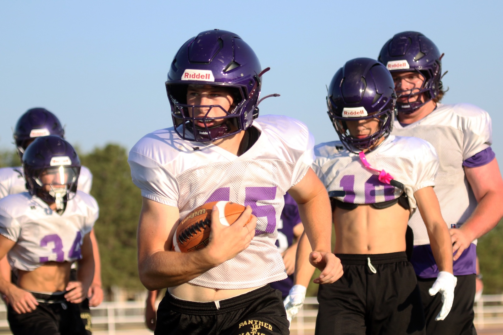 Young football players in purple helmets run on a sunny field, with the central player gripping a ball and wearing number 15.
