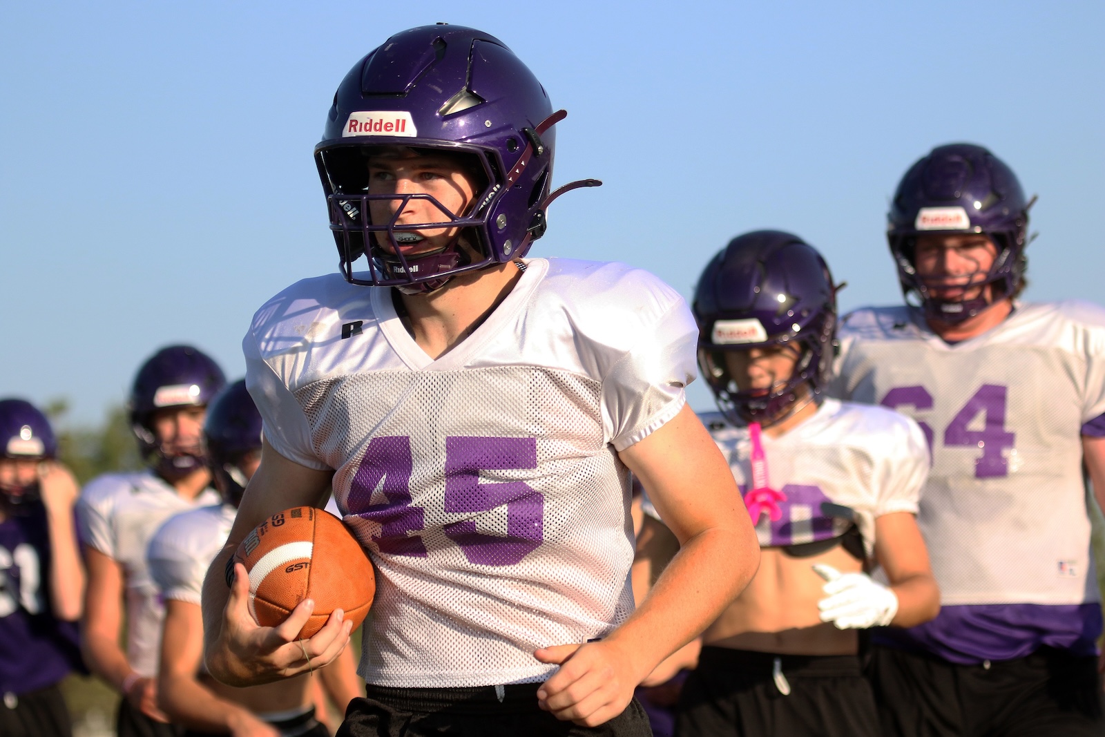 Football player wearing a purple helmet and white jersey with the number 45 runs with the ball in his right hand, teammates behind him on a sunny field