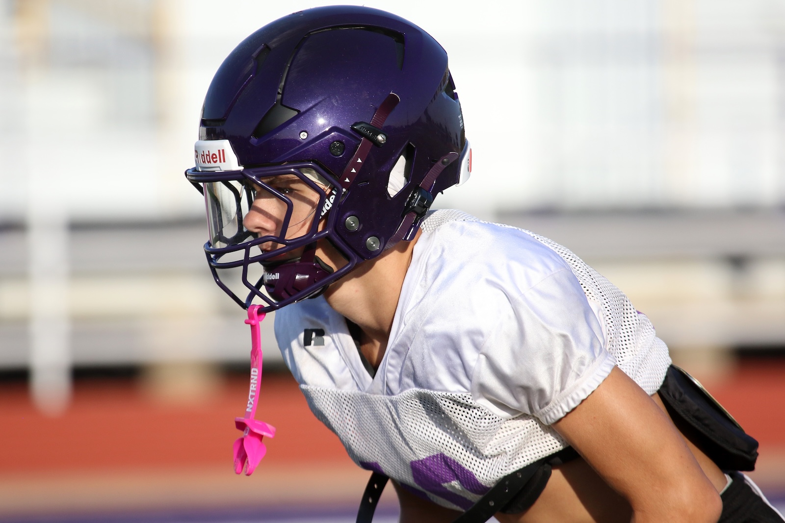 American football player in a purple helmet and white jersey crouches in a ready stance on the field.