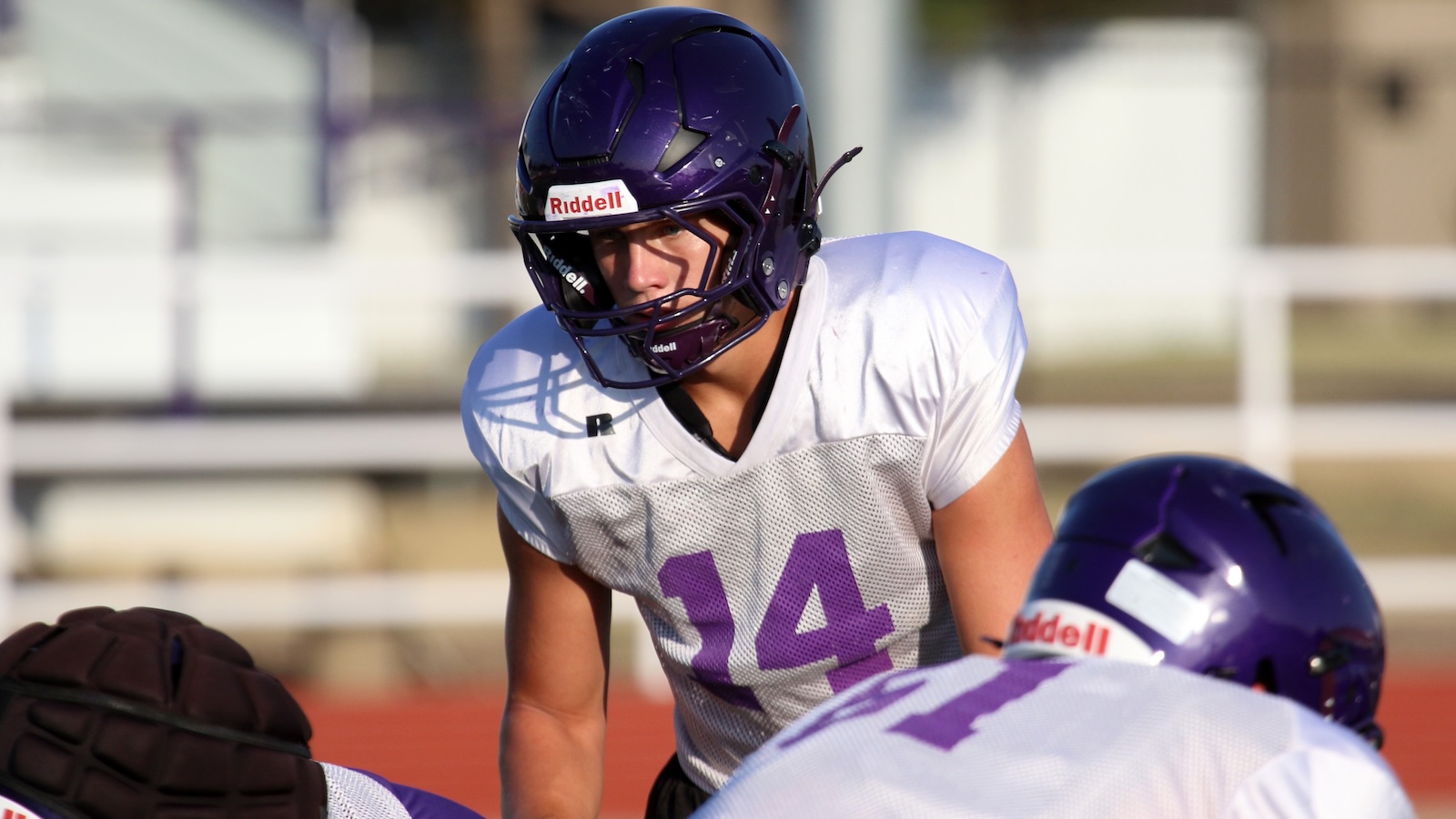 Football players in purple helmets warming up; foreground player wearing a white mesh jersey with purple number 14 ready for a snap.