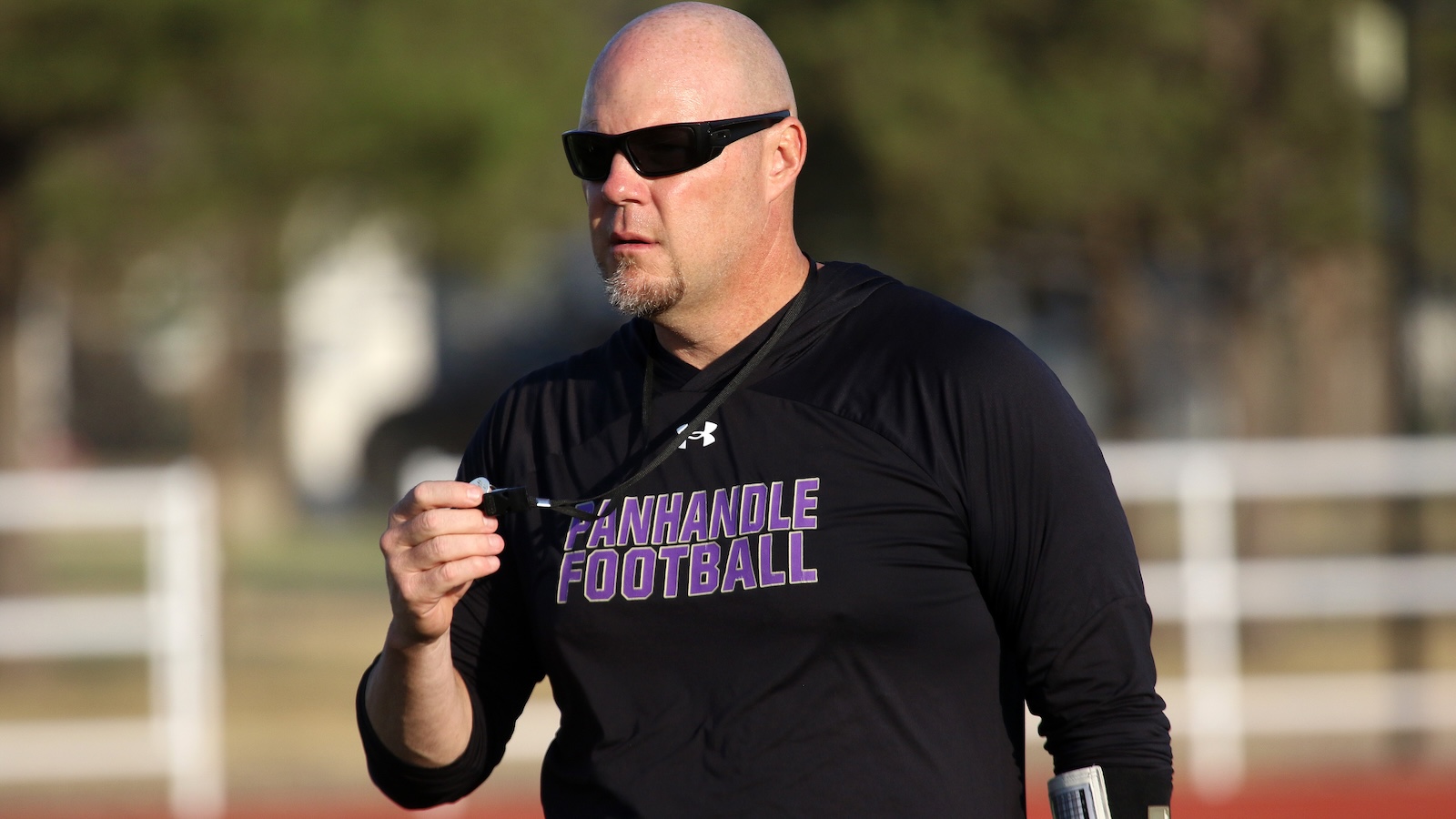 Male football coach wearing black shirt with'FANHANDLE FOOTBALL', sunglasses, and a whistle in hand on a field