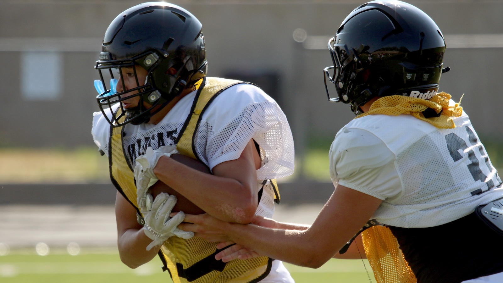 Two football players in helmets and pads practice on a field; one clutches a ball while a teammate grabs at his waist in a drill