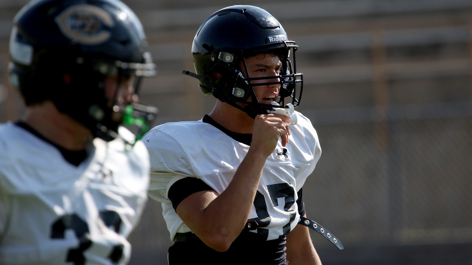 Football player in a white jersey and black helmet blows a whistle during practice on a field.