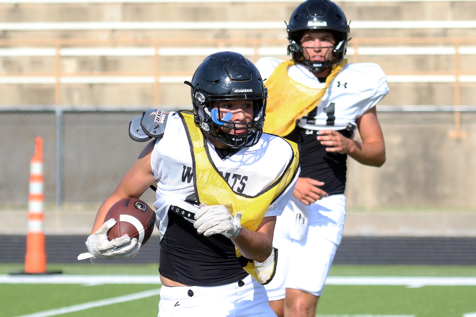 Football player in white jersey and yellow protective vest sprints with ball during practice; another player stands in the background.