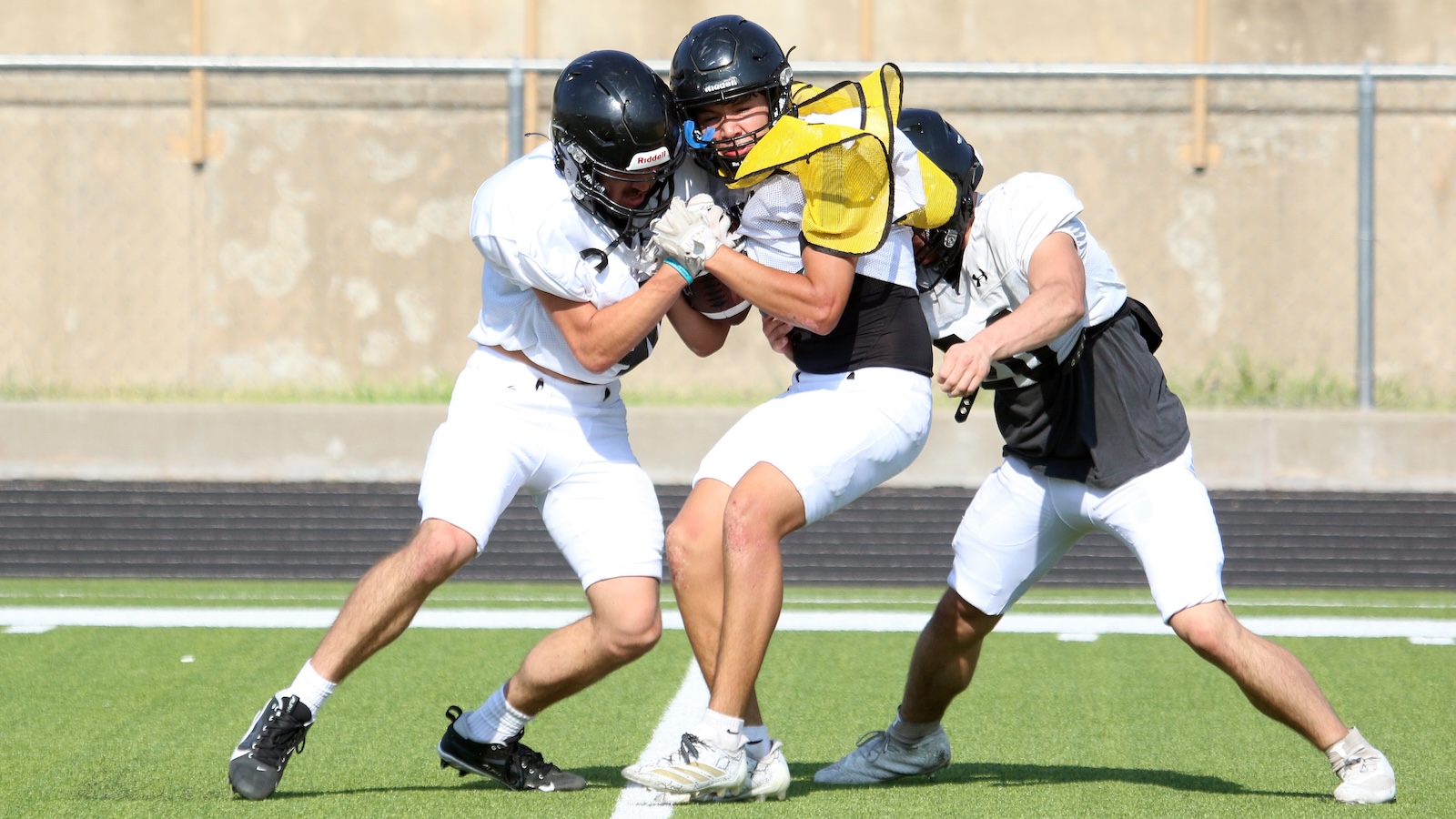 Three football players collide in a drill on a green field; a central player in a yellow protective vest is tackled by two white-uniform teammates.