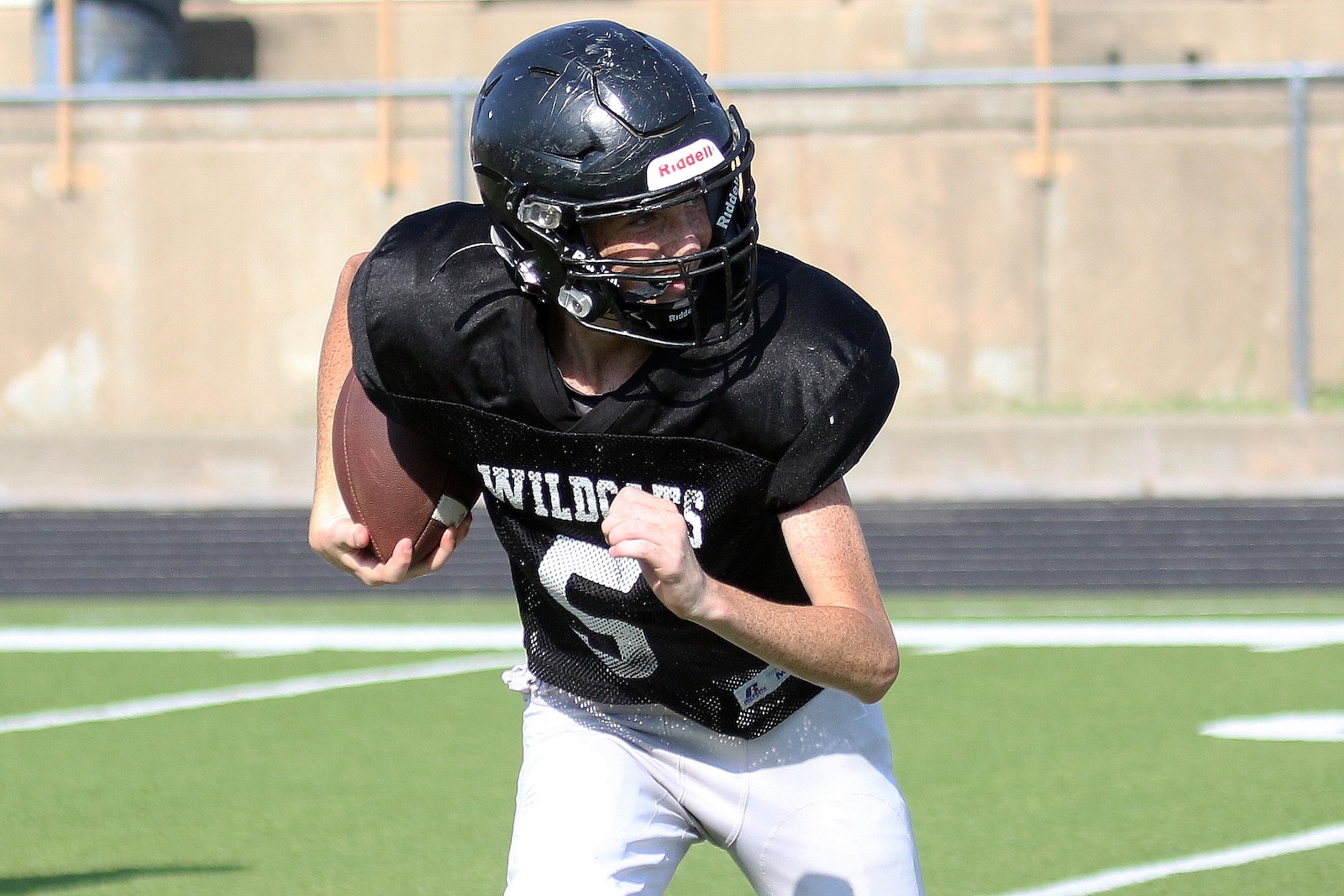 Football player in a black jersey and helmet carries the ball while running on a turf field.