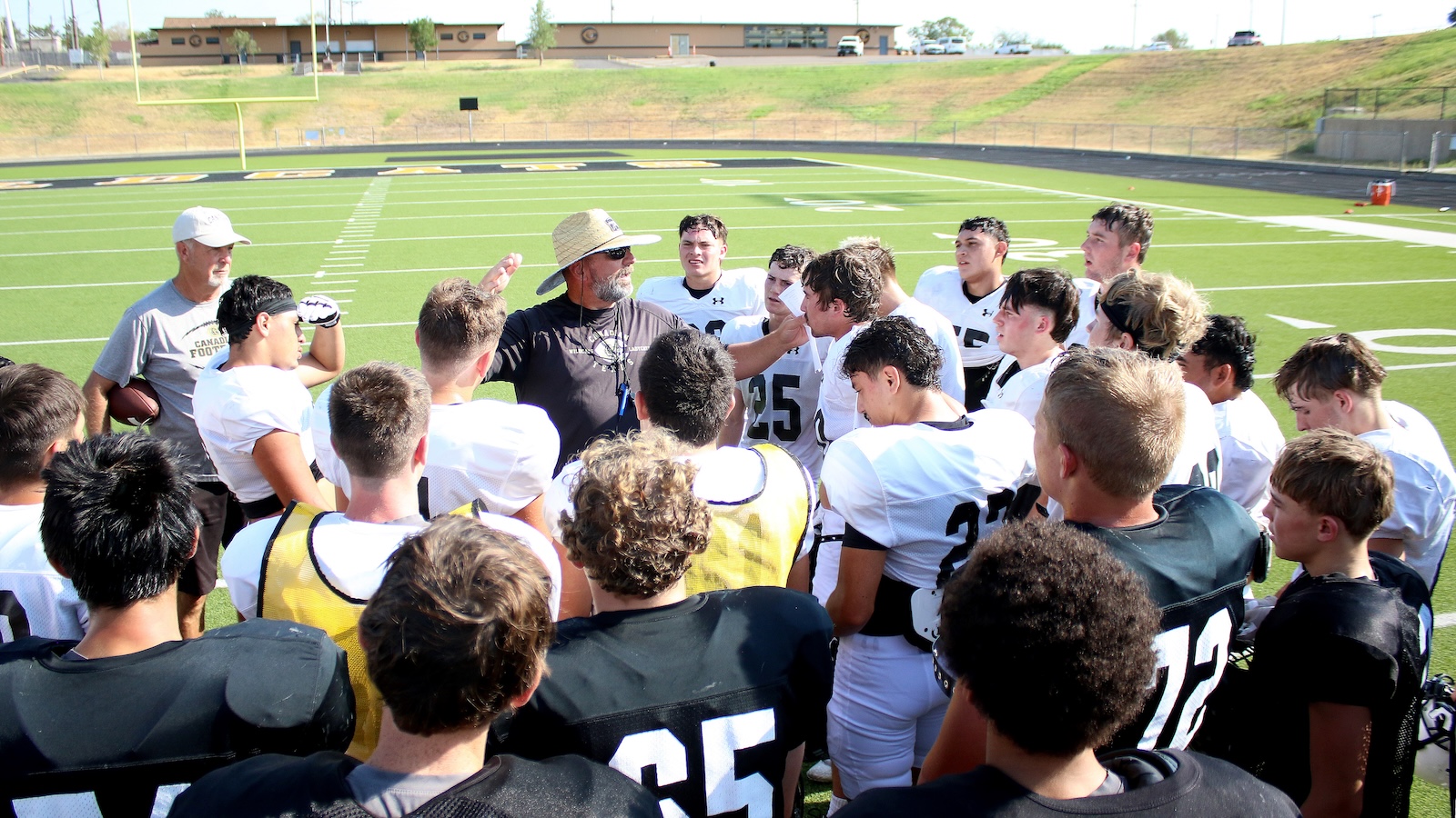 Football coach addresses a huddle of players on a sunny practice field, white uniforms and helmets nearby, turf field in background.