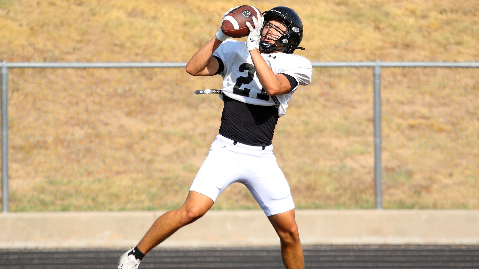 Football player in white and black uniform catches a football midair on a track field.