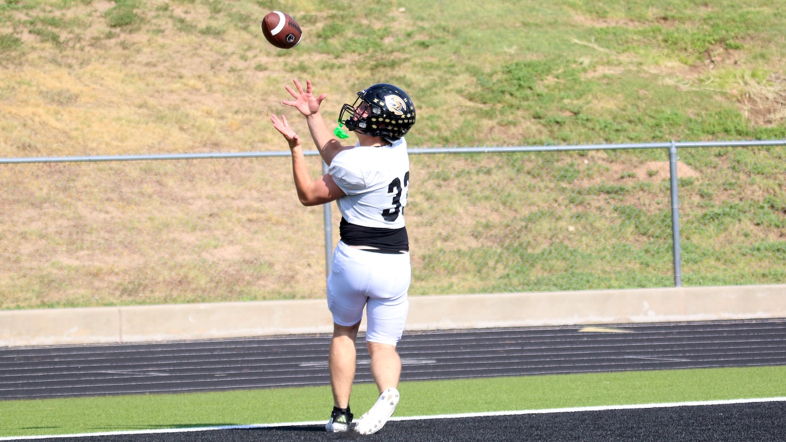 American football player in a white jersey leaps to catch a football on a track field, helmet on, number 34 visible behind him.
