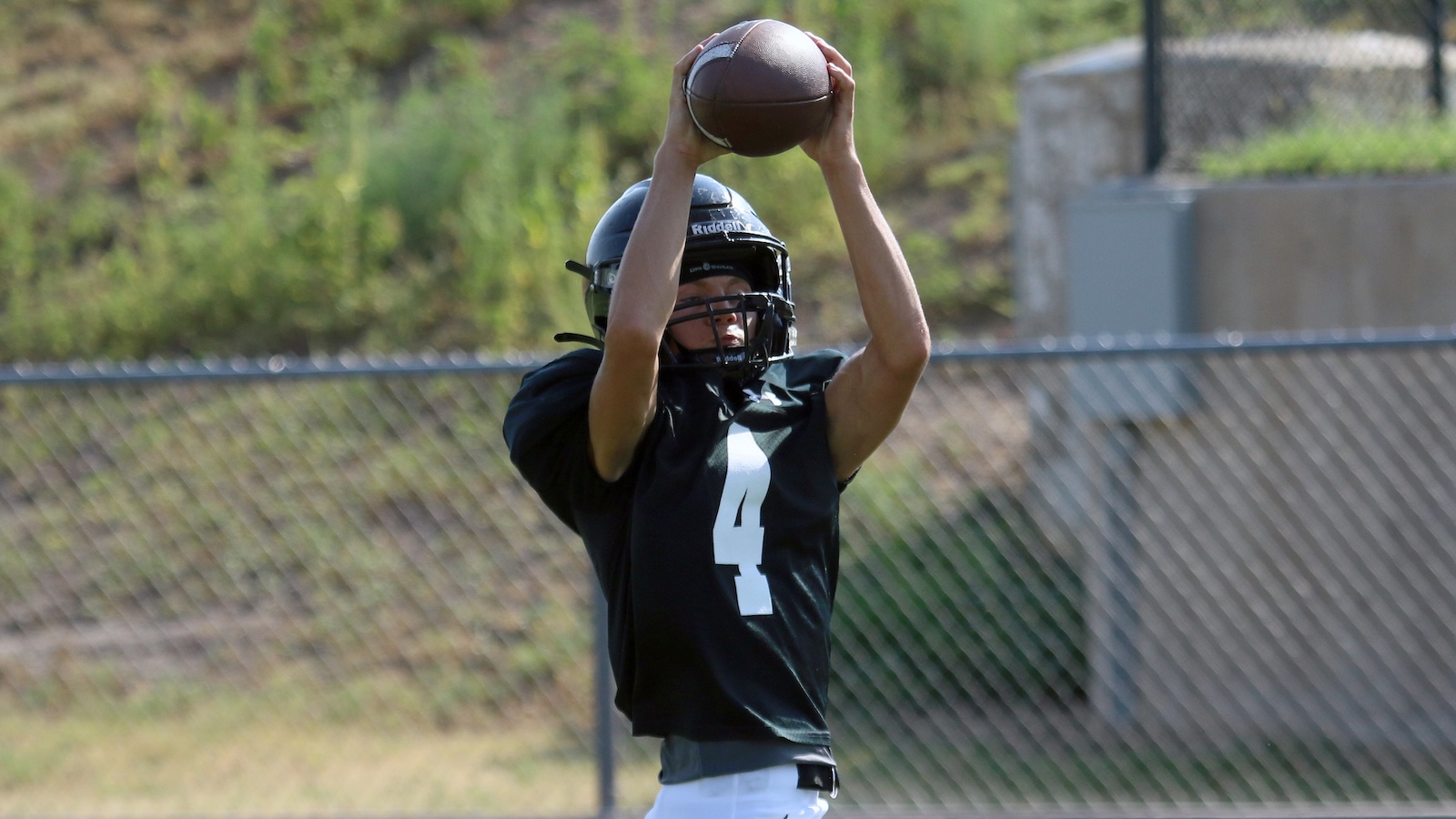 Football player in a black jersey (number 4) reaching up to catch a football on a field behind a chain-link fence.