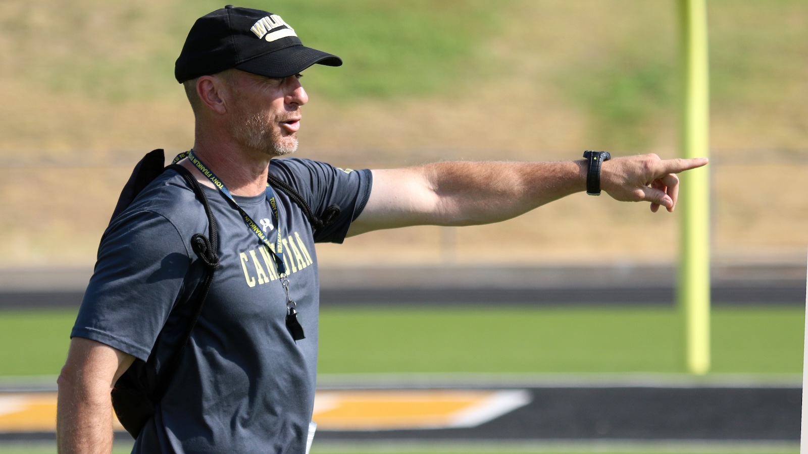 Coach on a track field in a gray athletic shirt and cap, pointing to the right with his arm extended and a whistle hanging from a lanyard