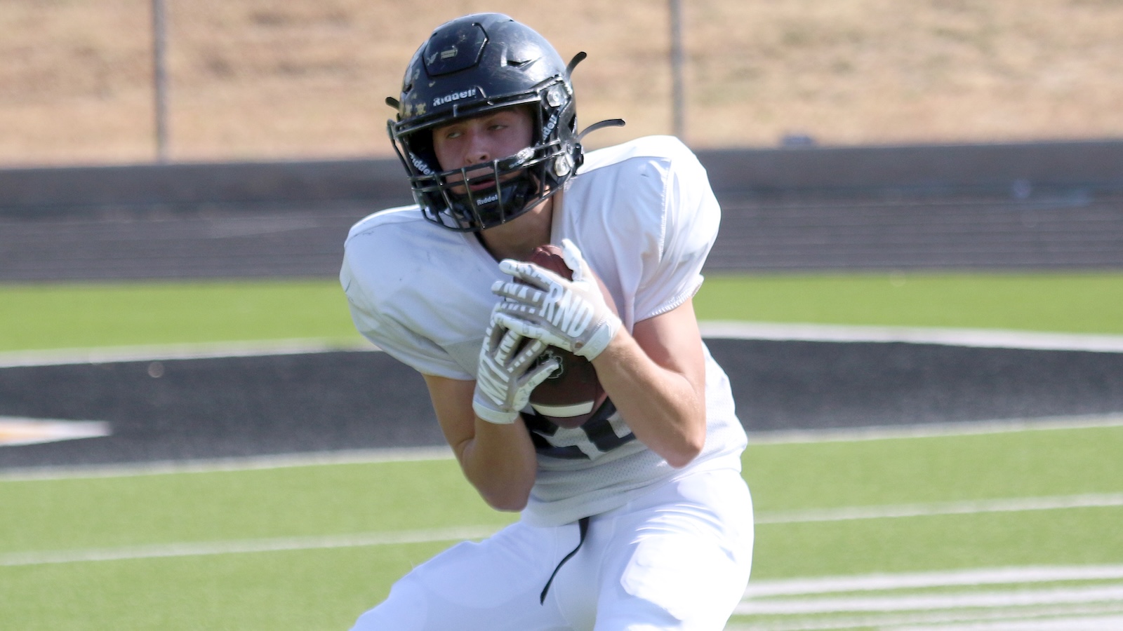American football player in a white uniform and black helmet runs with the football tucked under his arm on a green field.