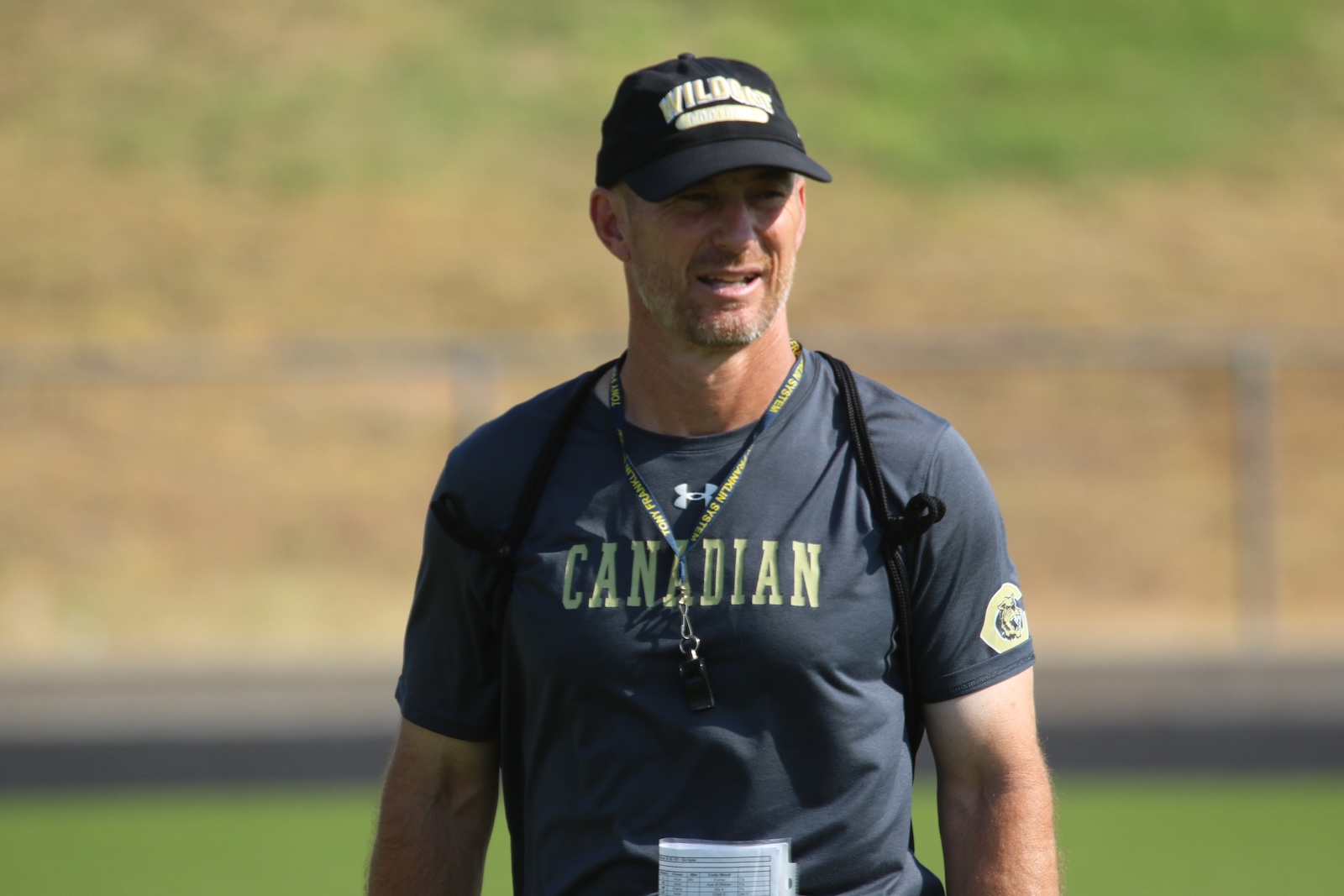 Man wearing a dark athletic shirt with'CANADIAN' across the chest, cap, and lanyard with a whistle, standing outdoors on a field.