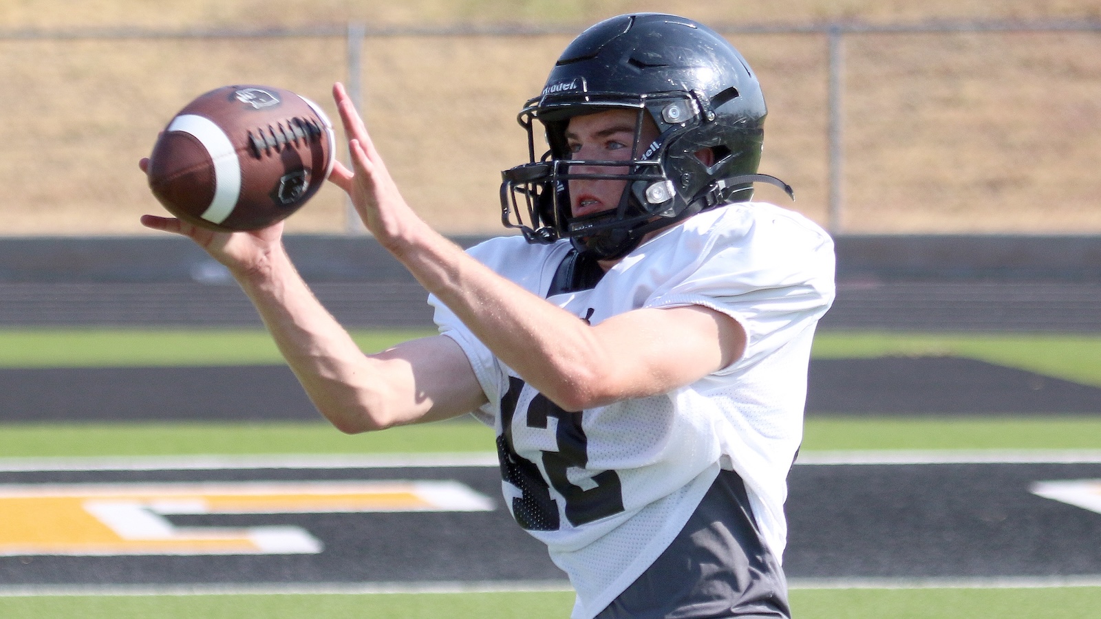 Football player in a white jersey and black helmet catches a football mid-air on a field.