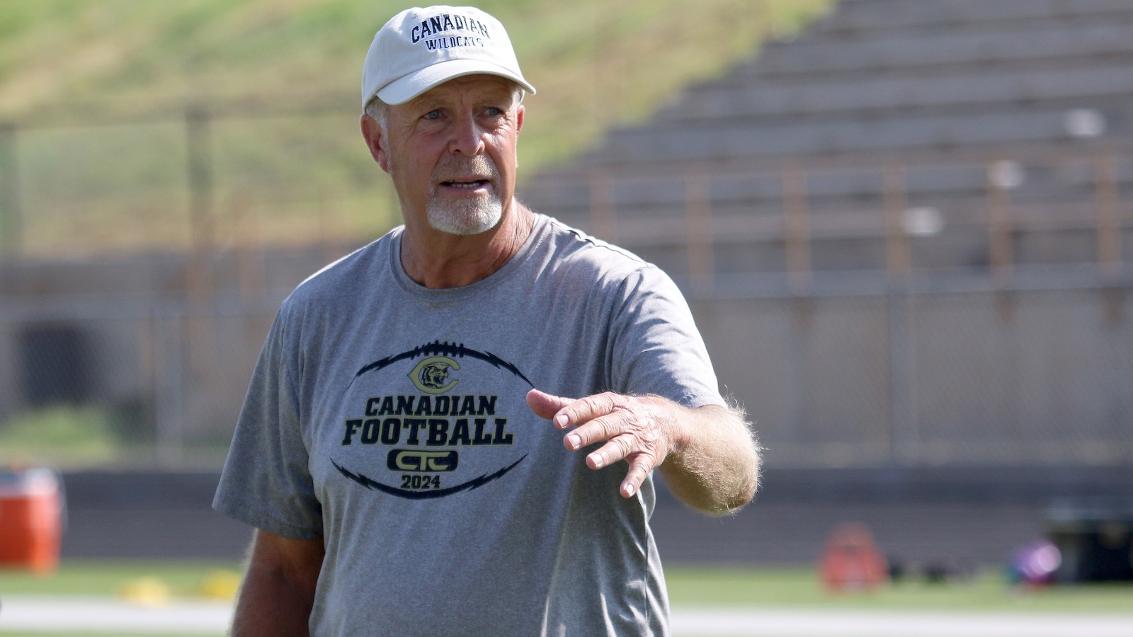 Older man in a gray Canadian Football T-shirt and white cap points as he speaks on a football practice field.