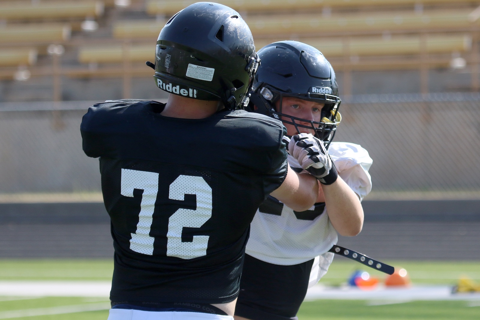 Two football players clash in a drill on a sunny field; one wears a black #12 jersey and a helmet.