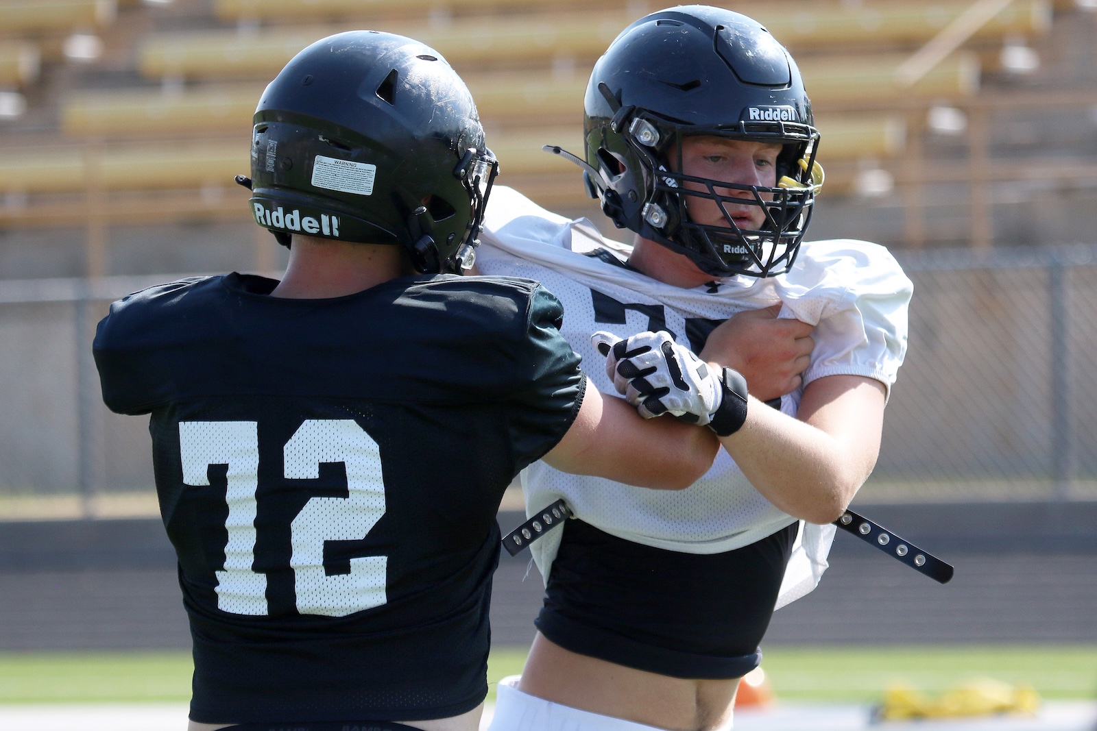 Two football players in helmets lock arms during practice on a field, one in black #72 jersey facing the other in white jersey.